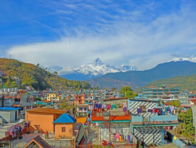 Cityscape of colorful rooftops with laundry hanging, set against a backdrop of snow-capped mountains under a clear blue sky.