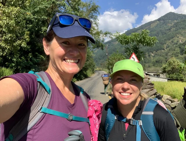 Two women wearing backpacks and hats smile for a selfie on a sunny day. A road, green hills, trees, and a cloudy sky are in the background.