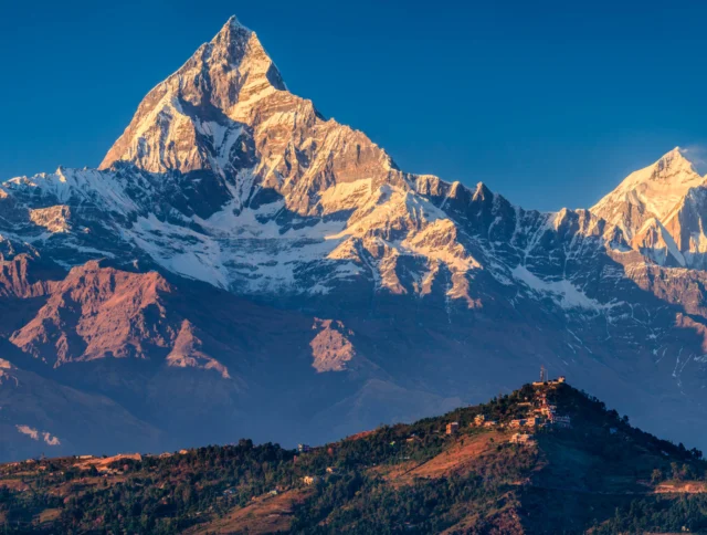 A majestic snow-capped mountain peak with rugged terrain and a clear blue sky. A hillside with scattered buildings is seen in the foreground.