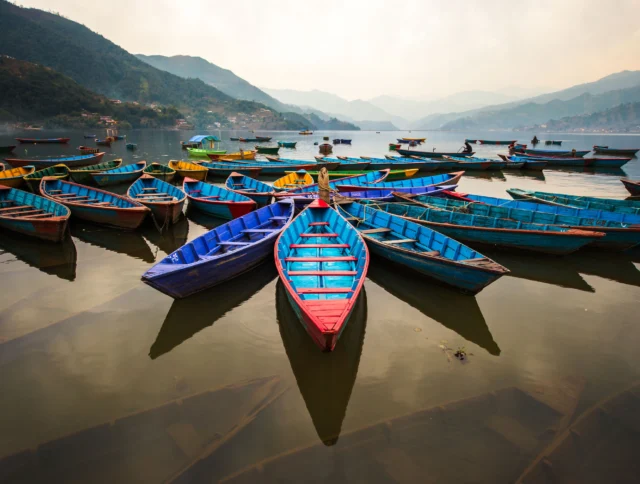 Colorful wooden boats are moored on a calm lake with mountains in the background under a cloudy sky.