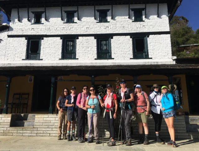 A group of hikers in outdoor gear stands in front of a two-story white building with dark green shutters.