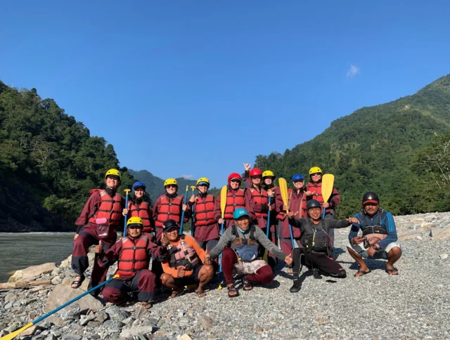 A group of people in life jackets and helmets stand by a river, holding paddles, with forested hills in the background.