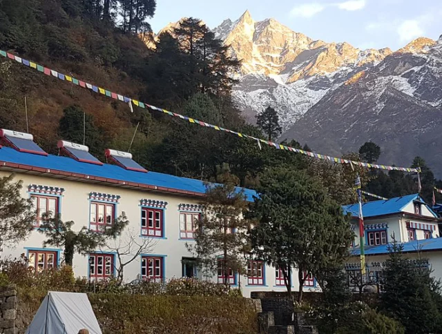 White and blue building with red windows, surrounded by trees, beneath snow-capped mountains. Prayer flags hang overhead.