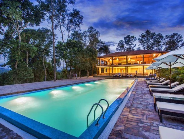 Outdoor pool area with lounge chairs and umbrellas, adjacent to a two-story building. Tall trees surround the area under a dusk sky.