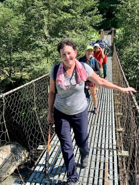 A group of people walking across a narrow rope bridge in a forested area, with one person at the front holding a walking stick.