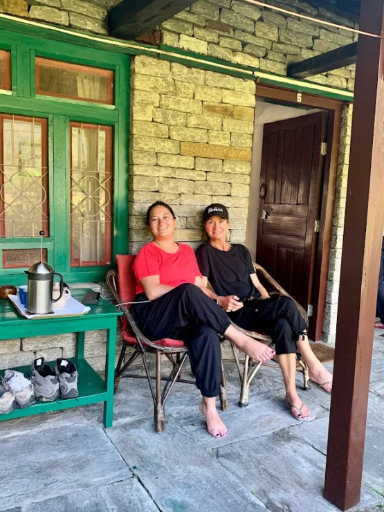 Two people sitting on chairs on a stone porch, smiling. They are dressed casually in black pants and tops. A small table with a thermos and shoes are visible nearby.