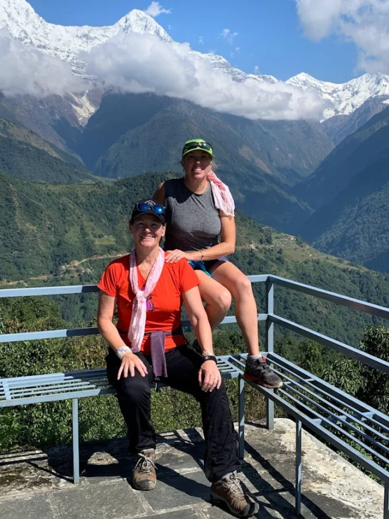 Two individuals sitting on a bench with a scenic background of snow-capped mountains and green valleys on a sunny day.