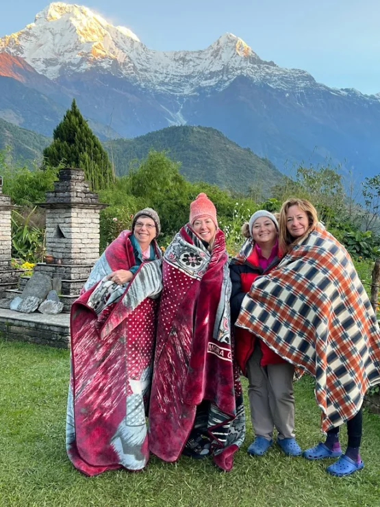 Four people wrapped in blankets stand outdoors in front of snow-capped mountains and greenery.