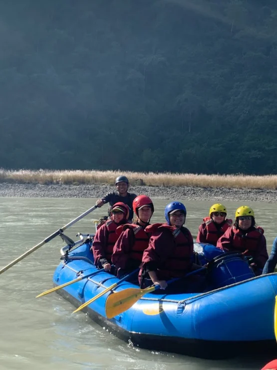 A group of people in life jackets and helmets are rafting on a calm river in a blue inflatable boat, with a forested background.