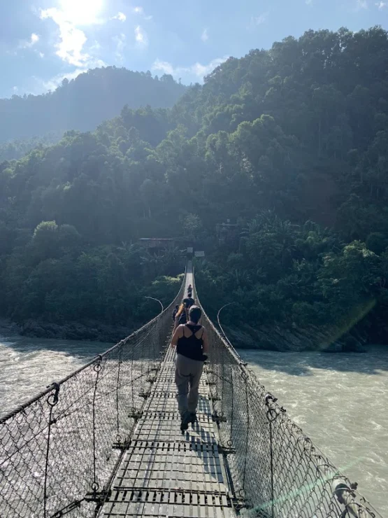 A person walks across a narrow suspension bridge over a river with forested hills in the background.