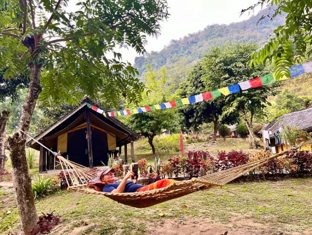 Person relaxing on a hammock outdoors in front of a wooden cabin, surrounded by trees and colorful flags, with hills in the background.