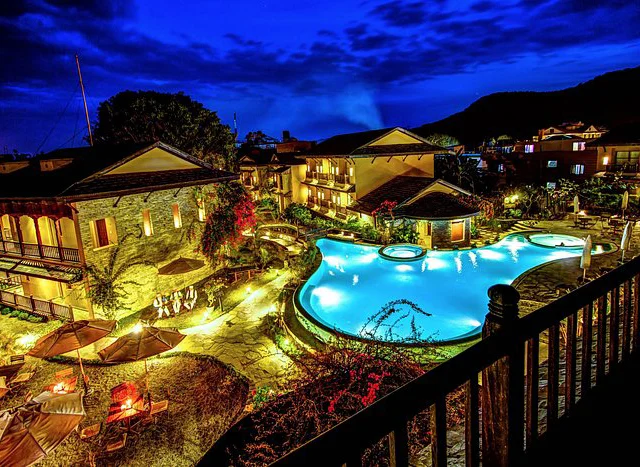 A night view of a brightly lit outdoor pool surrounded by buildings and trees. The scene includes lounge chairs and umbrellas, with a vibrant blue sky overhead.