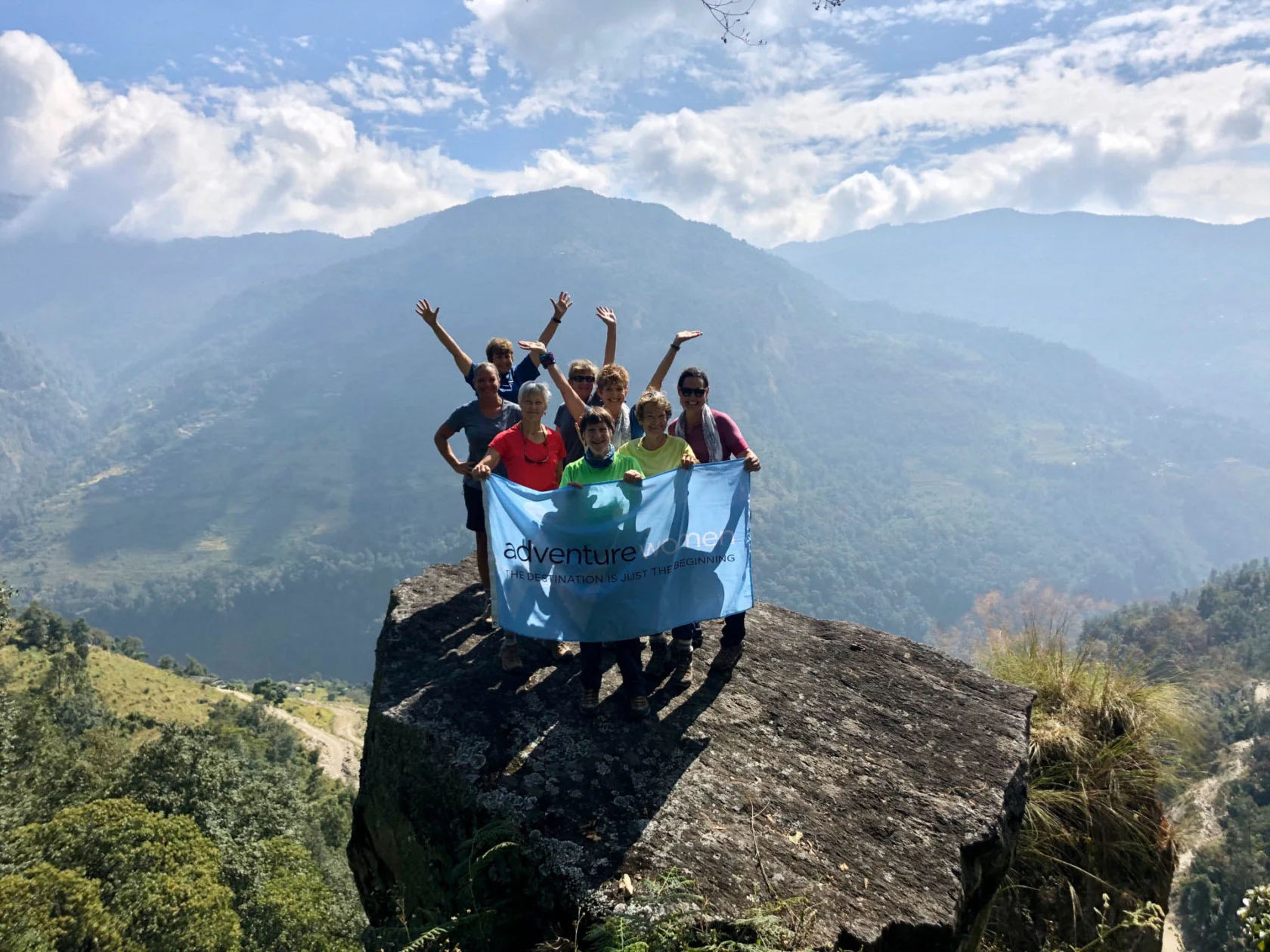 A group of people holding a banner stands on a large rock with a mountainous landscape in the background.