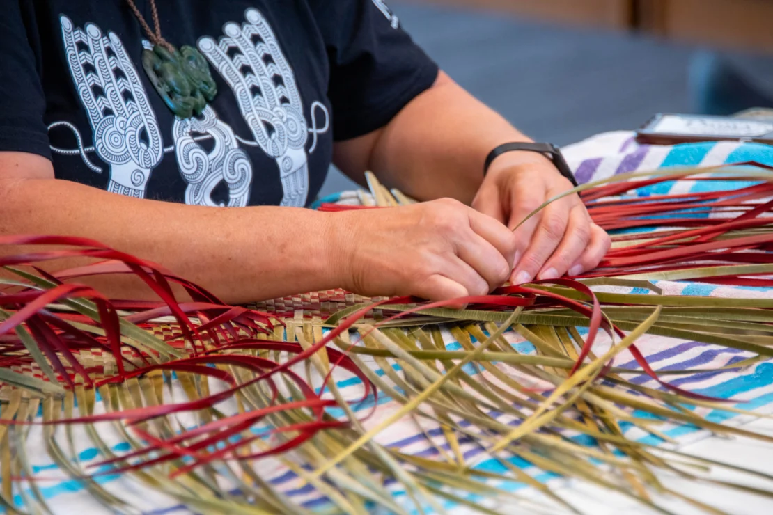 A person weaving red and green plant fibers on a striped tablecloth, wearing a black shirt with white designs.