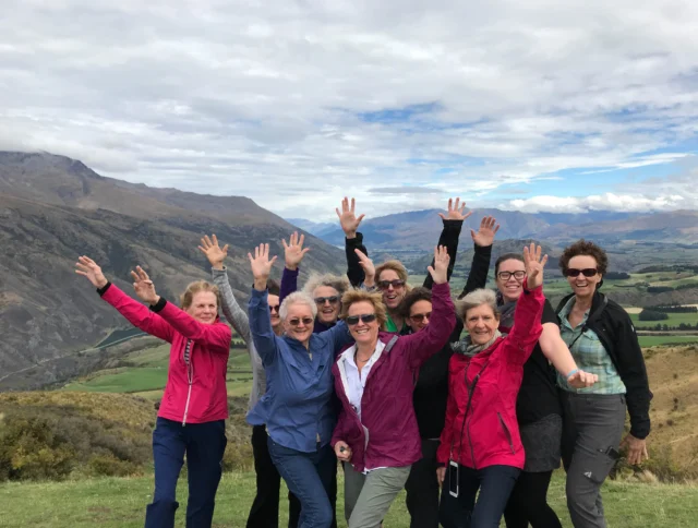 A group of people stand on a grassy hillside with mountains in the background, raising their hands and smiling at the camera.