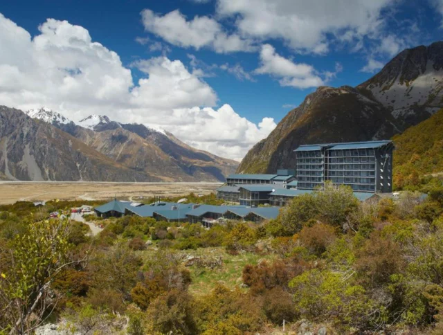 A modern hotel complex is nestled among lush green hills and mountains under a partly cloudy blue sky.