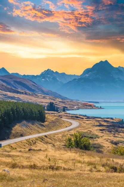 A winding road leads through grassy fields towards mountains at sunset, with a lake on the right and a colorful sky overhead.