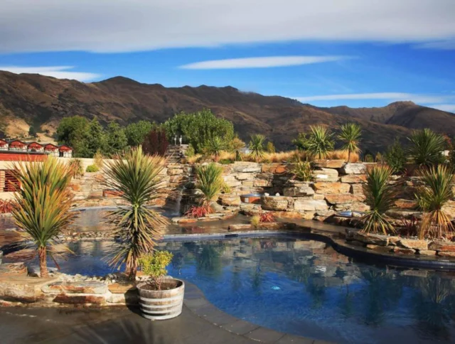 Outdoor pool surrounded by stone walls and potted plants, set against a backdrop of mountains and a blue sky with clouds.