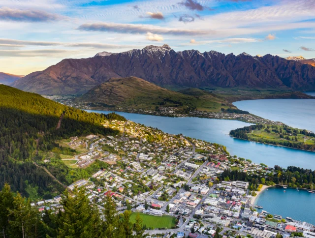 Aerial view of a town by a lake, with mountains in the background and a forested area in the foreground, under a partly cloudy sky.