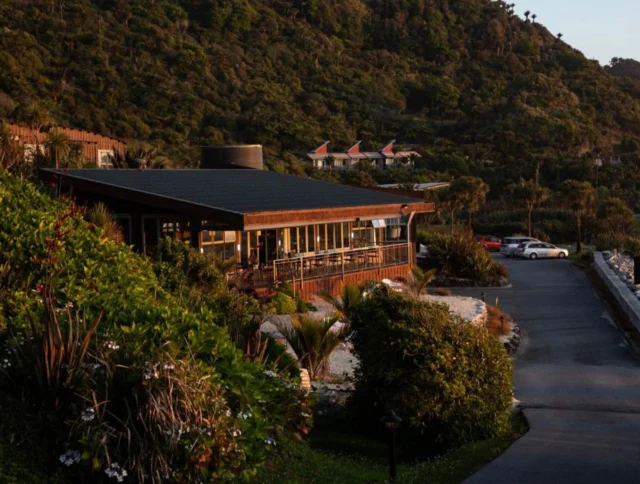 A wooden building with large windows sits surrounded by lush greenery, near a parking area, with a background of forested hills in the evening light.