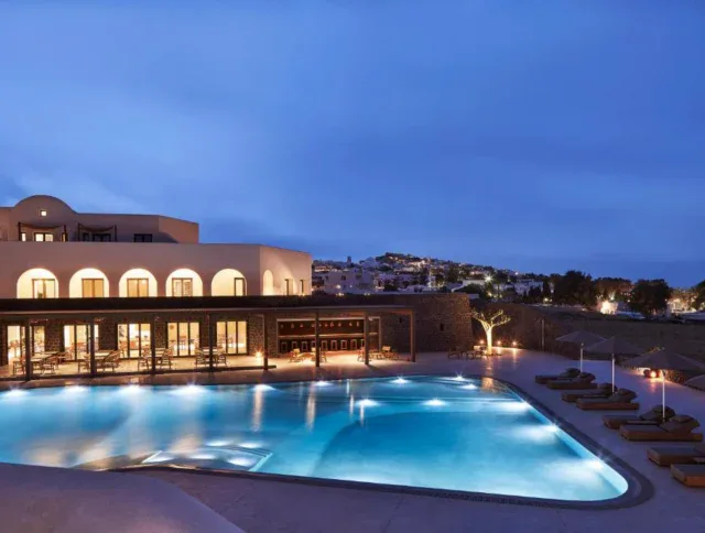 A modern hotel building with large windows overlooks a glowing outdoor swimming pool at dusk, surrounded by lounge chairs and umbrellas, with a hilly town in the background.