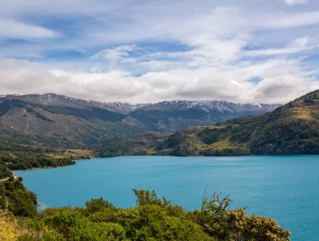 A serene turquoise lake surrounded by lush green hills and snow-capped mountains under a partly cloudy sky.