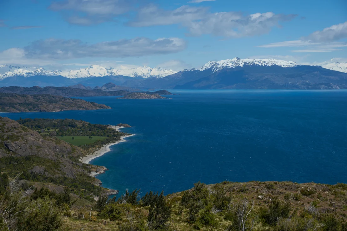 A panoramic view of a coastline with blue water, mountainous terrain, and snow-capped peaks under a partly cloudy sky.