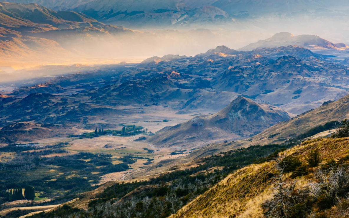 Aerial view of a vast, rugged mountainous landscape at sunset, with a soft golden mist hovering over the valleys and peaks.