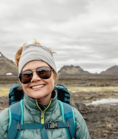 A person wearing sunglasses and a teal jacket smiles in a mountainous landscape with patches of snow.