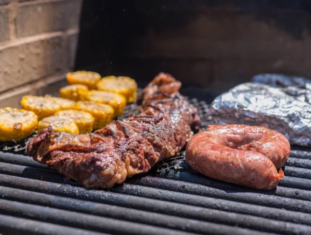 Grilled meat, sausage, corn slices, and foil-wrapped food cooking on a barbecue grill.