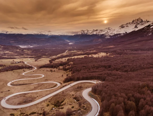 A winding road snakes through a vast landscape with snow-capped mountains in the background under a cloudy sky.