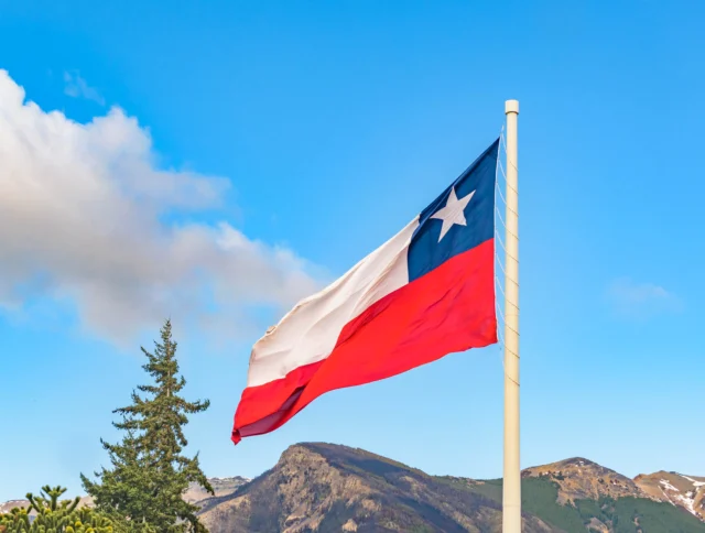 Chilean flag waving on a pole against a backdrop of mountains and a partly cloudy sky.