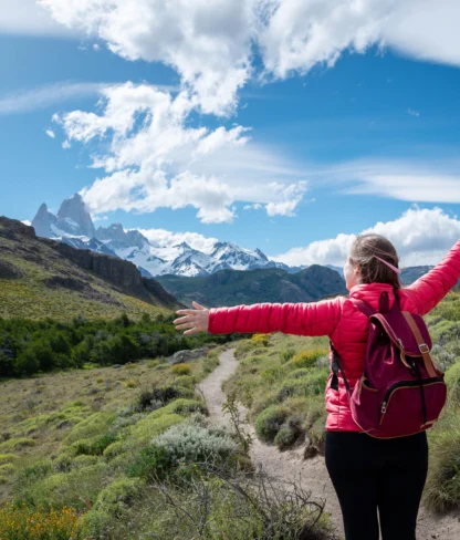 Person in a pink jacket and backpack stands on a trail with arms outstretched, facing snowy mountains under a partly cloudy sky.