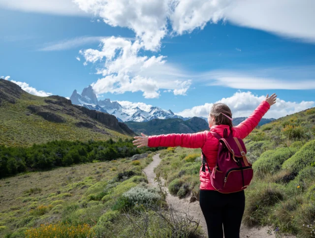 Person in a pink jacket and backpack stands on a trail with arms outstretched, facing snowy mountains under a partly cloudy sky.