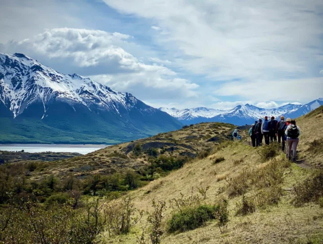 A group of hikers walks along a grassy trail with snow-capped mountains and a lake in the background under a cloudy sky.