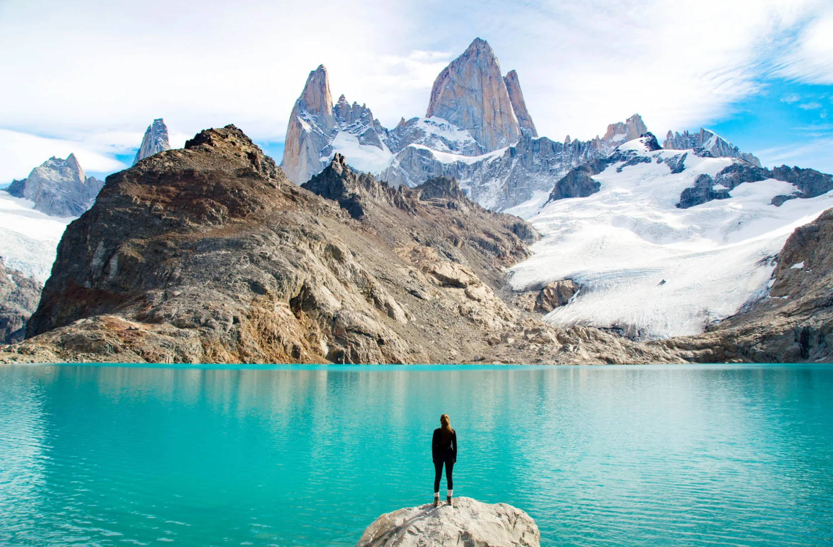 A person stands on a rock, looking at a turquoise lake with snowy mountains in the background under a partly cloudy sky.