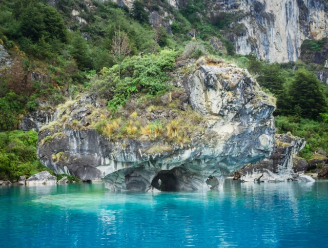 Rock formation with natural archway surrounded by clear turquoise water, set against a backdrop of lush greenery and rocky cliffs.