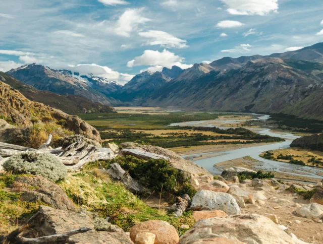 A scenic landscape with a winding river, lush valley, and distant mountains under a partly cloudy sky. Stones and greenery are in the foreground.