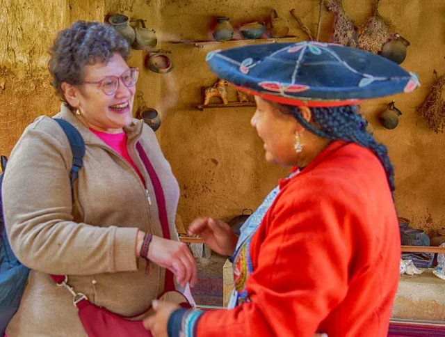 Two women, one in casual clothing and one in traditional attire, converse and smile in a rustic room with pottery and dried herbs hanging on the wall.