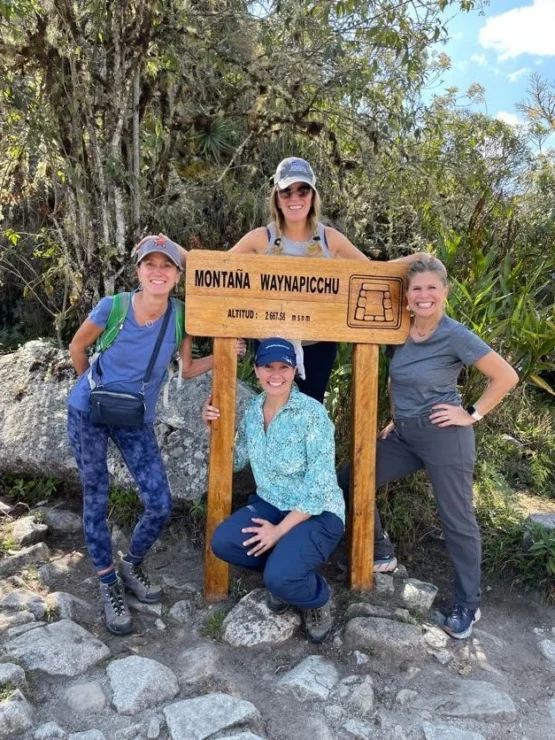 Four people pose by a wooden sign for Montaña Waynapicchu, showing altitude 2,667 meters, surrounded by trees and rocks.