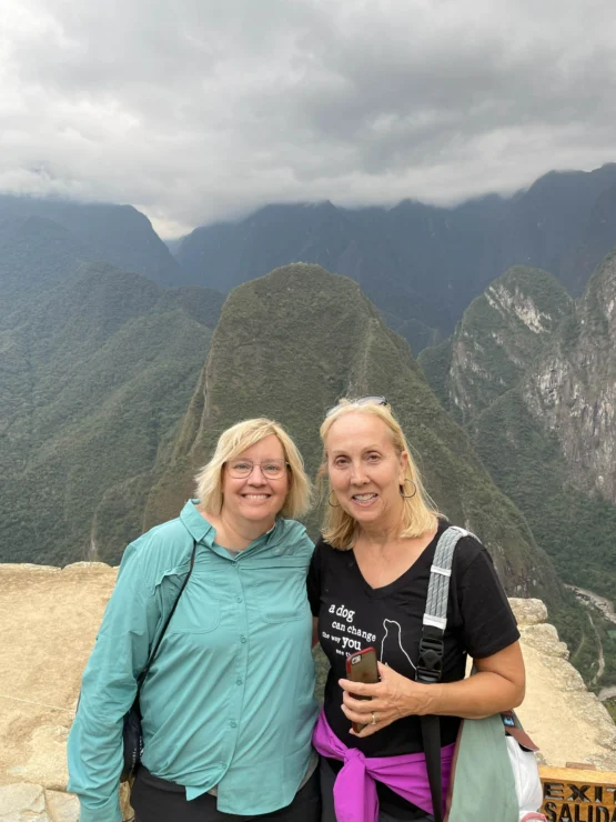 Two women smiling in front of a mountainous landscape with cloudy skies. They are casually dressed and standing close to each other.