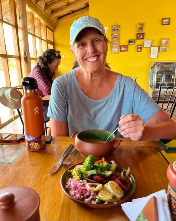 A person in a blue shirt and cap smiles while holding a spoon over a clay bowl at a table with a plate of colorful food. A water bottle is on the table; another person is in the background.