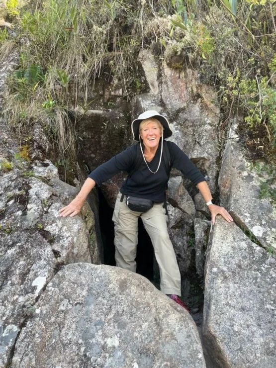 A person wearing outdoor clothing and a hat stands in a rocky crevice surrounded by greenery.