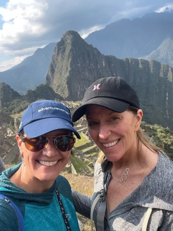 Two people in casual outdoor attire smile for a selfie with Machu Picchu's iconic mountain landscape in the background.