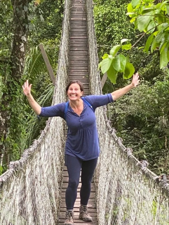 Person smiling and posing on a narrow rope bridge in a lush, green forest.