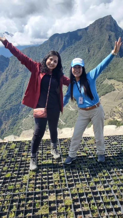 Two people pose with outstretched arms on a scenic mountain overlook, with lush green hills and a cloudy sky in the background.