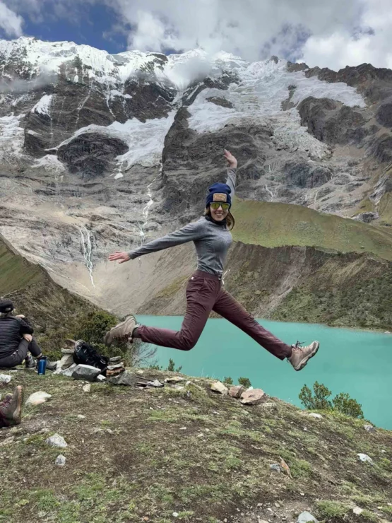 Person jumps mid-air with arms and legs outstretched in front of a turquoise lake and snow-capped mountains. Backpackers sit nearby.