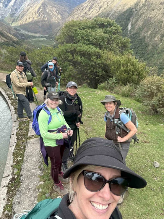 A group of hikers posing on a trail with mountainous scenery in the background. They're wearing backpacks and outdoor gear, smiling at the camera.