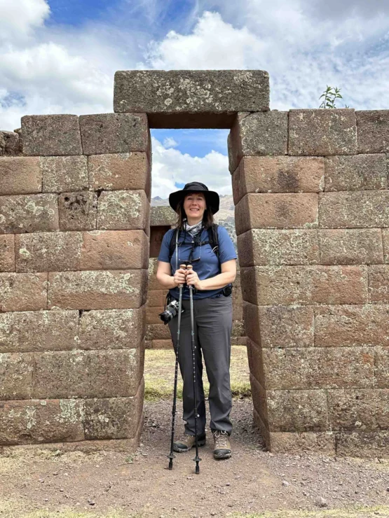 A person stands in a stone doorway, holding hiking poles and wearing a hat, backpack, and camera. The sky is partly cloudy.