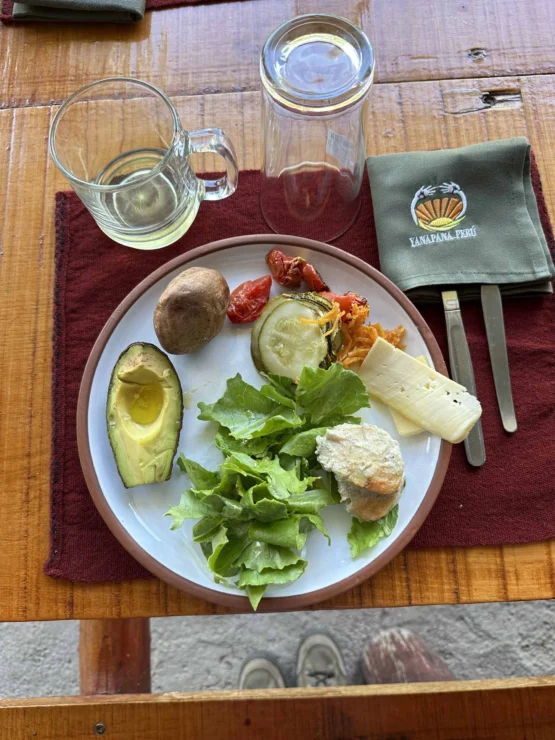 A plate with avocado, lettuce, a potato, grilled vegetables, cheese, and bread. A glass and mug are next to it on the table.